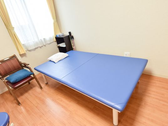青いベッドが１台設置されており、白い枕が置かれている。ベッドの左側に椅子があり、座面に青いクッションが載せられている。