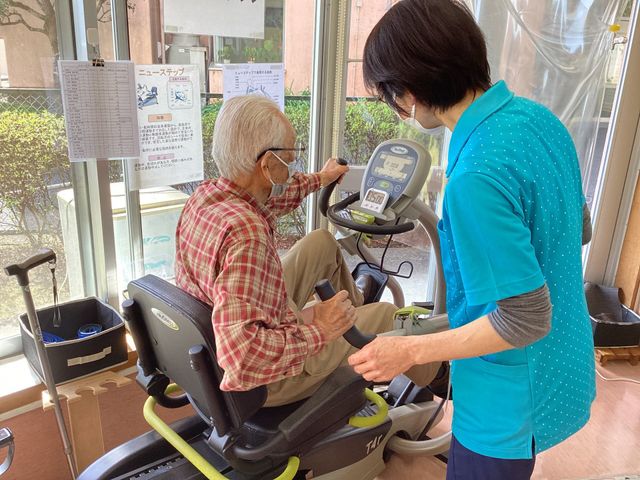 運動器具を使用する様子 機能訓練室 スタッフの支援を受けながら、運動器具を使った体力維持に取り組む様子です。利用者様の安全と健康を第一に考えた空間が整っています。