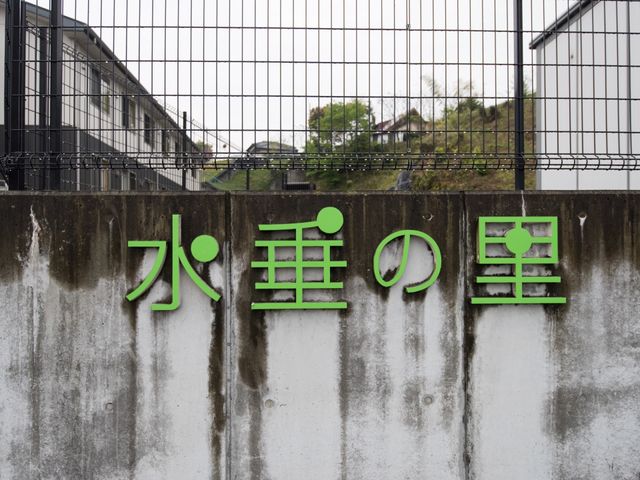 緑の看板の風景 施設名 緑色が鮮やかな看板が設置された門扉です。あなたの新たな職場への第一歩を、ここから始めませんか。