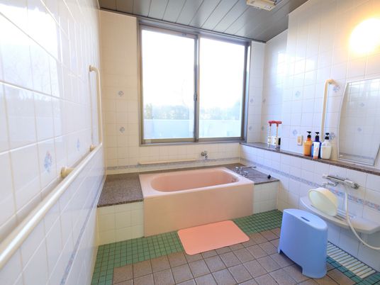 数種類のタイル張りになっている浴室で、床には滑り止めマットが敷かれている。ピンクの浴槽が設置され、大きな窓から外光が差し込んでいる。