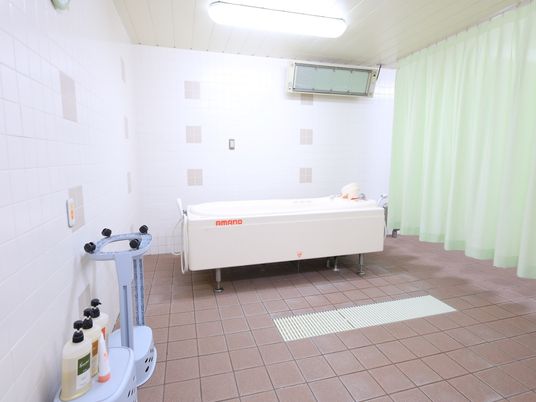 奥の壁際に寝位入浴用の浴槽が置かれている。右側に緑のカーテンが引かれている。左の壁に非常ボタンが取りつけられている。