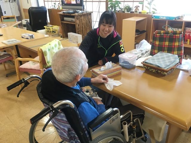 笑顔のスタッフと住人が会話する様子のリビング リビング 明るい空間で、スタッフが入居者と親しげに交流しています。温かみのあるリビングでの和やかな時間です。