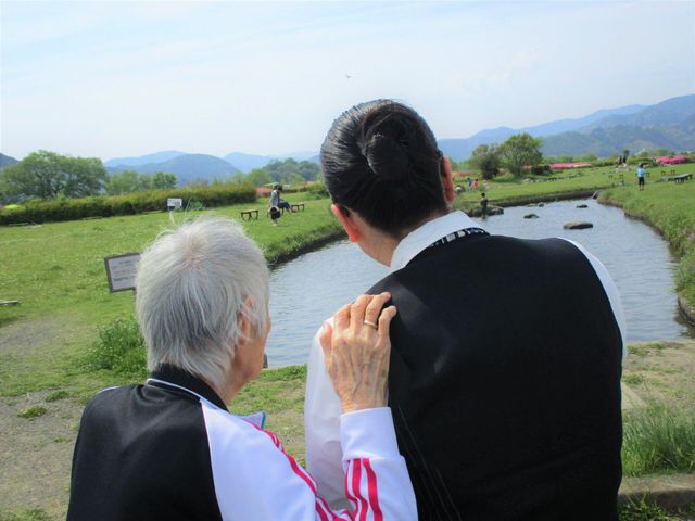 自然を楽しむ高齢者と介護者 リハビリ 緑豊かな中庭で、ゆったりと流れる川を見つめる高齢者の方々。介護者の優しいサポートが心地良いひと時を約束します。