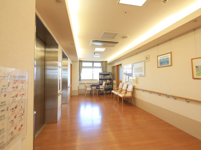 廊下の明るい光景 エレベーターホール 床の木目調とナチュラルな色合いが特徴の廊下です。手すりや座席も設置され、落ち着いた雰囲気の中で休憩を取れるスペースがあります。