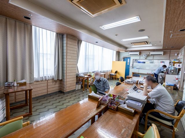 広々とした食堂の様子 ダイニングルーム 光が差し込む落ち着いた雰囲気の食堂で、利用者が新聞を読んだり、スタッフが作業をしています。フラットな床で移動が容易な設計が特徴です。