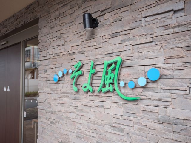 石壁に施設名の看板 玄関 屋外に設置された緑色の文字が印象的な施設の名前の看板です。これから就業を検討している方には、新たな職場への第一歩を感じていただけるかもしれません。