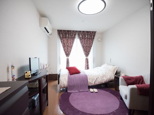 施設の写真 白を基調とした明るい居室である。エアコンや介護用ベッド、液晶テレビ、タンス、ソファが完備されている。