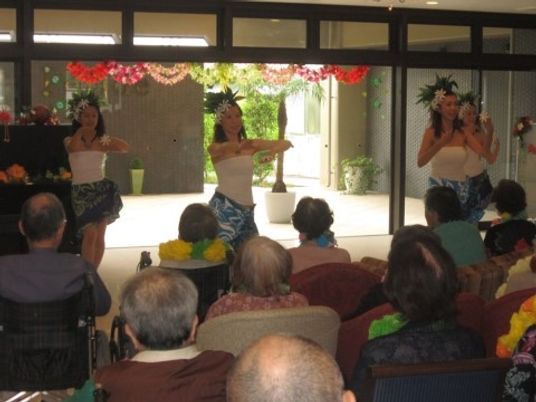 ４人のフラダンスの演者が踊りを踊っている。多くの入居者様が椅子や車椅子に座って観賞している場面である。