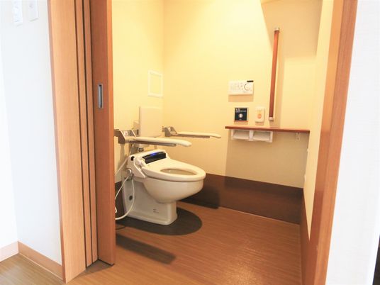 施設の写真 居室のトイレには引き戸がついている。水タンクのない便器で、壁に設置されたボタンで水を流すようになっている。