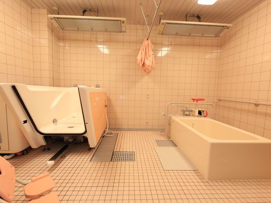 施設の写真 浴室に、通常の浴槽と介護浴槽が、間を空けて設置されている。その間は、ピンク色のカーテンで仕切ることができる。