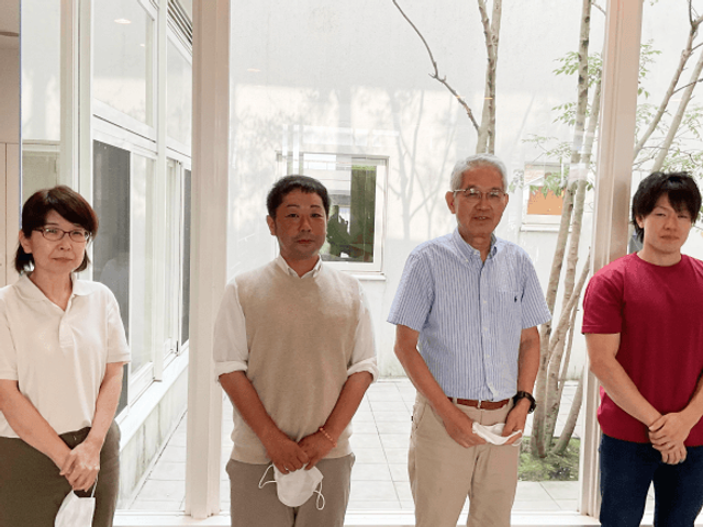 職員が並ぶ中庭 中庭   白い壁に囲まれた明るい中庭で、職員の方が4人並んで立っています。自然光が入りやすく、落ち着いた雰囲気が伝わります。