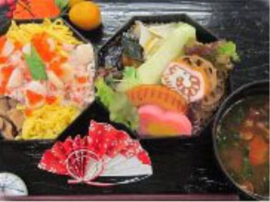 施設の写真 八角形の重箱に入れられたお食事。メインのちらし寿司の他に汁物も用意されている。お弁当の周りに可愛らしい装飾がされている。