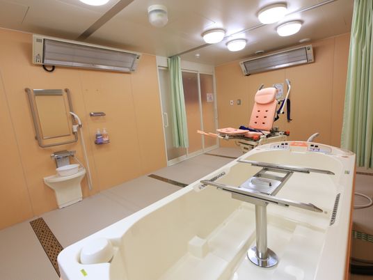 施設の写真 広い浴室で、特殊寝台が置いてあっても、洗い場には十分なスペースがある。シャワーの隣には、備え付けの棚がある。