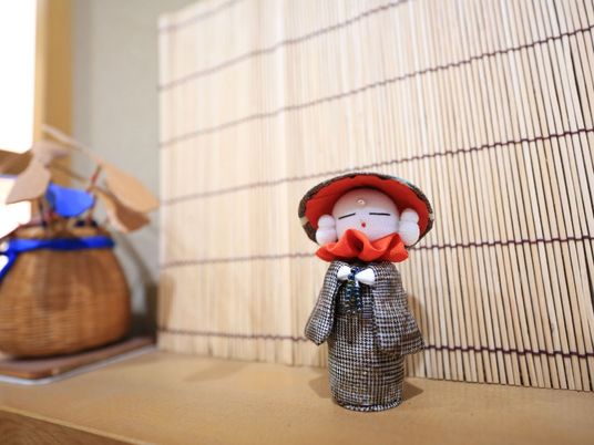 木製の棚の上に置かれた、布製の可愛らしいお地蔵様の人形。背後にはすだれがあり、側のかごには竹とんぼが入っている。