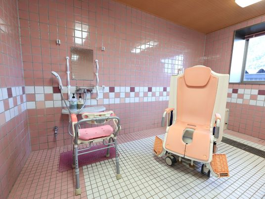 施設の写真 ピンク色のタイルに囲まれた、窓のある広い浴室。洗い場には体を固定して使うことができる椅子と、入浴用の車椅子が置かれている。
