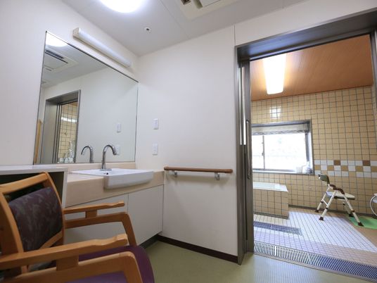 施設の写真 浴室へと続く脱衣所。壁に手すりと大きな鏡のついた洗面台が設置されている。ひじ掛け付きの椅子が置かれている。