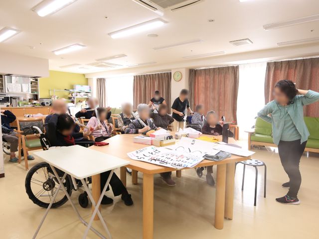 活動する利用者と職員 談話室 ゆとりのある食堂では、利用者が集い様々な活動を楽しんでいます。手作りのアイテムに囲まれ、職員のサポートを受けながら、心地よいひと時を過ごしている様子が伝わります。